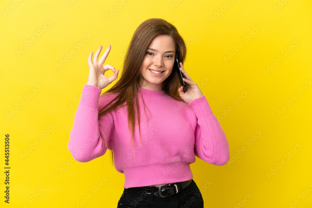 Teenager girl using mobile phone over isolated yellow background showing ok sign with fingers Teenager girl using mobile phone over isolated yellow background showing ok sign with fingers