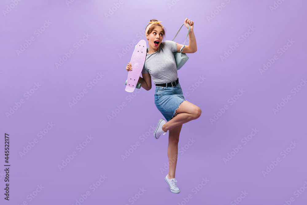 Full length shot of girl in denim skirt and shirt posing with longboard. Shy young woman in grey outfit and white modern sneakers looking into camera.. Full length shot of girl in denim skirt and shirt posing with longboard. Shy young woman in grey outfit and white modern sneakers looking into camera..