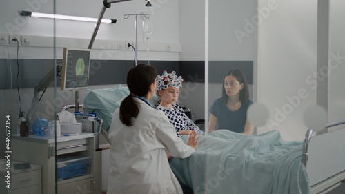 Sick girl child patient wearing eeg headset with sensors while pediatrician doctor monitoring brain tomography evolution checking expertise on monitor. Kid resting in bed in hospital ward