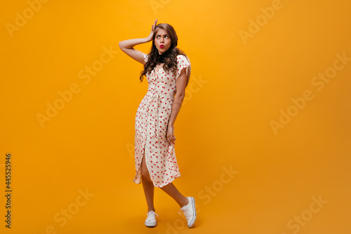 Charming woman in white dress mannerly posing on orange background. Modern young girl in cool clothes, hat, and sneakers looking up.. photo