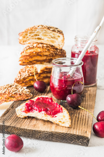 Homemade cherry jam, fresh cherries and cut lye rolls on a wooden board on a white table, vertical