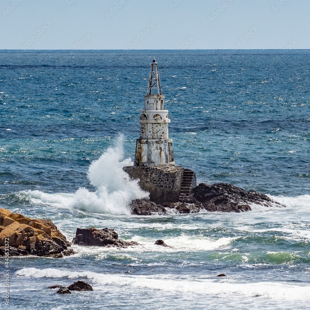 Attractive old lighthouse seascape, sea waves crashing into the rocks, sunny day. White splashing water drops rising high above the blue water surface. Southern Black Sea coast, Ahtopol, Bulgaria.