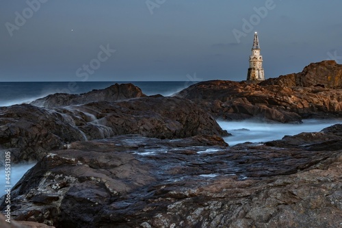 Attractive night seascape, sea waves splashing over rocks, shining lighthouse. Evening darkness, long exposure, blurry water trails and misty steam. White Southern Black Sea coast, Ahtopol, Bulgaria.