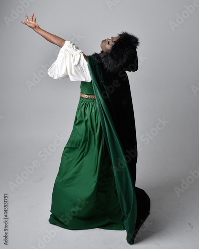 Full length portrait of pretty African woman wearing long green medieval fantasy gown and velvet cloak, sitting on a light grey studio background.