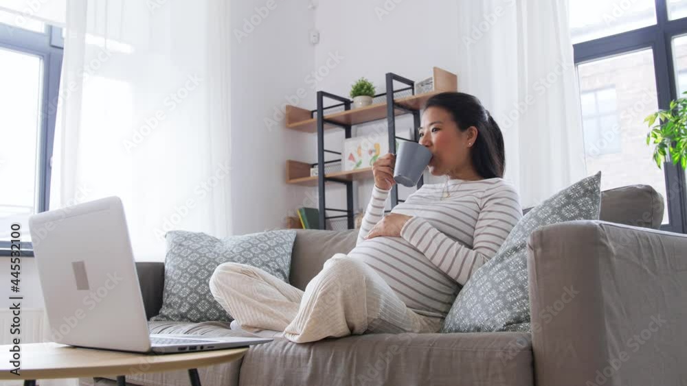 pregnancy, rest, people and expectation concept - happy smiling pregnant asian woman with laptop computer sitting on sofa at home and drinking tea pregnancy, rest, people and expectation concept - happy smiling pregnant asian woman with laptop computer sitting on sofa at home and drinking tea