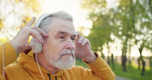 Close up of happy senior Caucasian handsome grey-haired bearded man standing outdoors alone putting headphones on and listening to music. Leisure concept, park, recreation, walk, sport photo