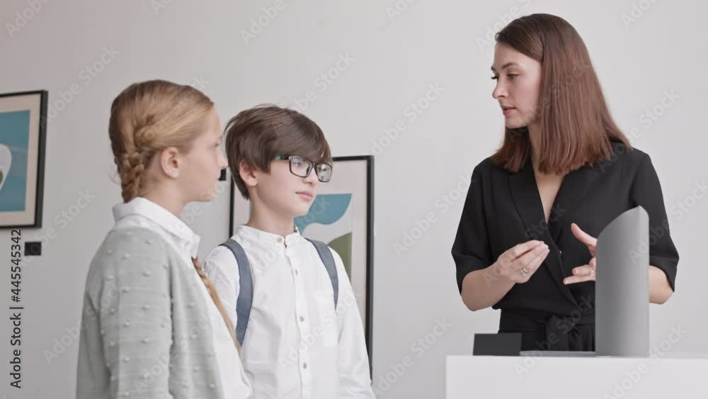 Medium shot of female Caucasian museum worker standing nearby art exhibit, talking to school students, smiling Medium shot of female Caucasian museum worker standing nearby art exhibit, talking to school students, smiling