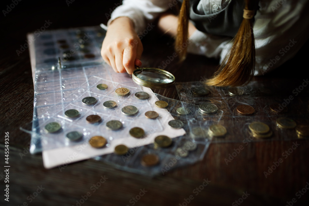 Cute cute european girl child in glasses with magnifying glass looks at coins, concept children and financial literacy, children and money, coin collecting and numismatics, dark style - obrazy, fototapety, plakaty 