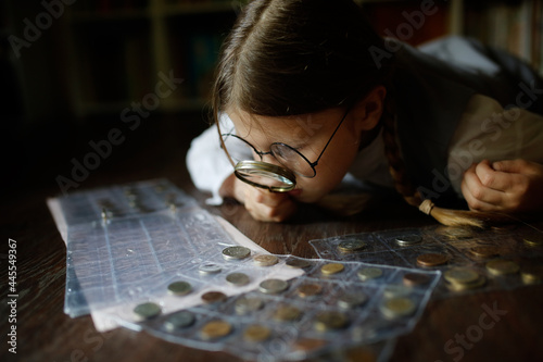 Cute cute european girl child in glasses with magnifying glass looks at coins, concept children and financial literacy, children and money, coin collecting and numismatics, dark style photo