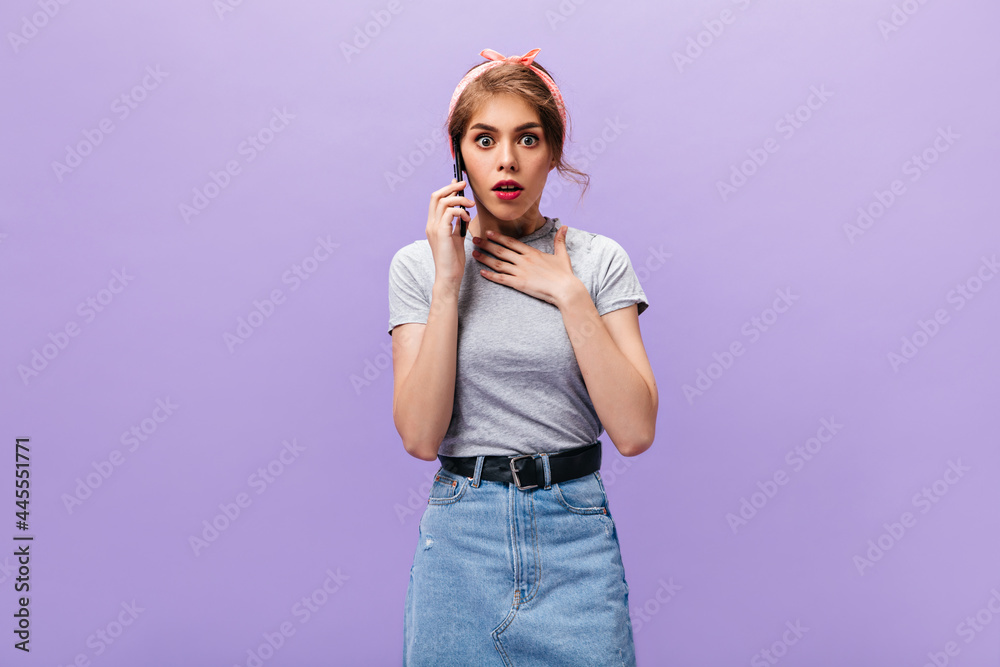 Woman looks shocked while talking on phone. Stylish girl in pink headband, grey t-shirt and light denim skirt posing on purple background.. Woman looks shocked while talking on phone. Stylish girl in pink headband, grey t-shirt and light denim skirt posing on purple background..
