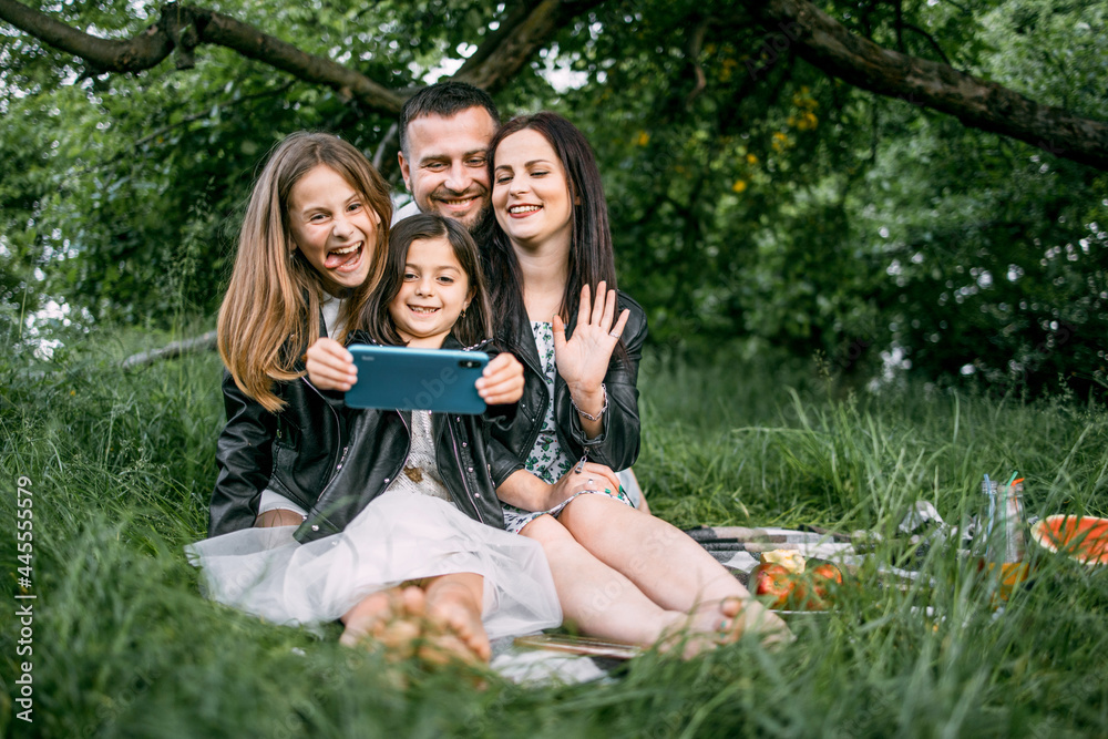 Fototapeta premium Positive family sitting together on soft plaid utdoors and taking selfie on modern smartphone. Young parents with two kids saving memories about happy summer picnic time.