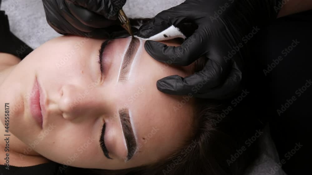 Master applies dark coloring pigment to correct shape of eyebrows with a special machine. The beautiful face of young girl model on sofa in a beauty salon before microblading, permanent makeup tattoo