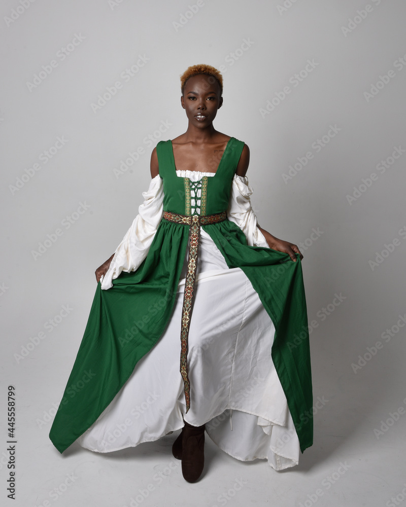 Full length portrait of pretty African woman wearing long green medieval fantasy gown, standing and dancing pose on a light grey studio background.