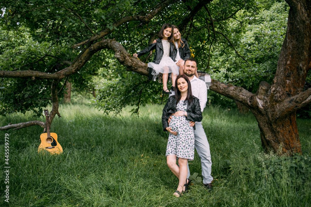 Fototapeta premium Two little sisters sitting on branch of old tree while father gently embracing pregnant mother. Caucasian family waiting for new addition.