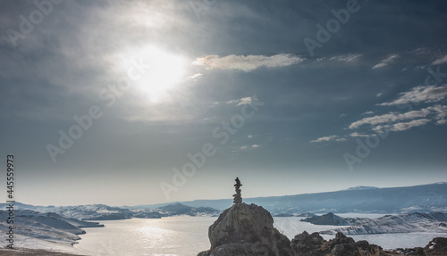 Winter Siberian Landscape. In the blue sky white clouds. Sun glare on the ice of a frozen lake. On the granite boulder there is a pyramid of stones. Snow on the hills. Baikal. Pastel shades
