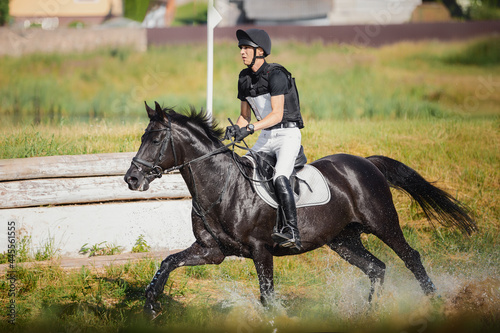 closeup portrait of rider man galloping fast in water to obstacle on black stallion horse during eventing cross country competition in summer