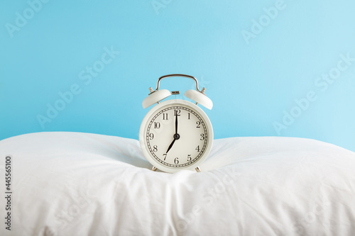 White alarm clock on pillow at light blue wall background. Pastel color. 7 o'clock in morning. Waking time concept. Closeup. Front view.