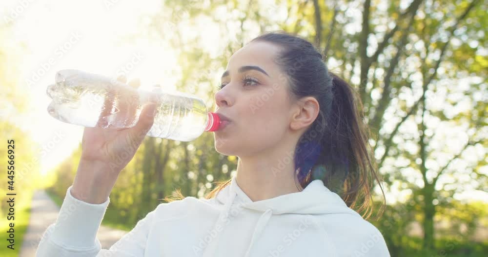 Close up portrait of Caucasian young beautiful female standing outdoors drinking water from bottle after workout on street in sun lights, Sporty lifestyle, cardio workout. Sportswoman concept Close up portrait of Caucasian young beautiful female standing outdoors drinking water from bottle after workout on street in sun lights, Sporty lifestyle, cardio workout. Sportswoman concept