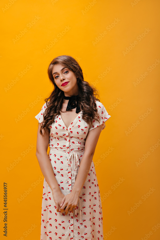 Curly woman in white dress smiling on orange background. Young shy girl with wavy hair in cherry print clothes looking into camera.. Curly woman in white dress smiling on orange background. Young shy girl with wavy hair in cherry print clothes looking into camera..