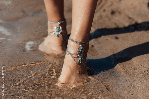 Close up of the feet in boho jewelry of a woman walking on the beach barefoot. Sunny summer day. Selective focus photo