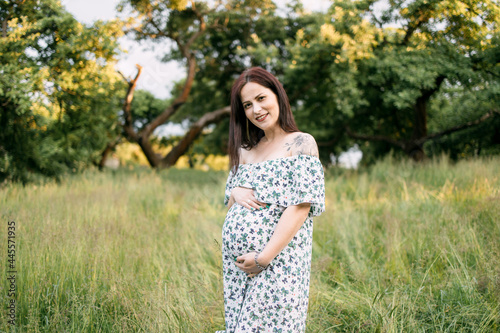 Fototapeta Naklejka Na Ścianę i Meble -  Charming young woman wearing summer dress with bare shoulder standing among high grass at green garden and genty embracing pregnant tummy. Concept of people, nature and expectation.