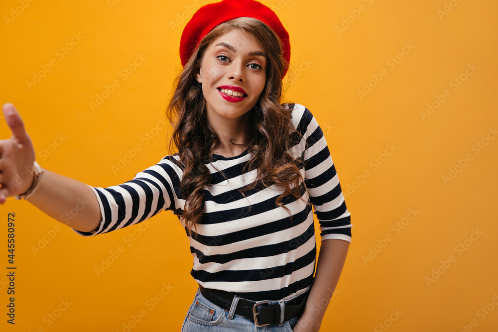 Attractive curly woman in beret makes selfie on orange background. Modern girl with wavy hair in striped sweater posing on isolated backdrop..