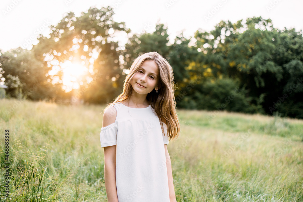Portrait of cute girl in white summer dress smiling and looking at camera while standing at green garden. Charming little lady with blond hair enjoying warm days outdoors. Portrait of cute girl in white summer dress smiling and looking at camera while standing at green garden. Charming little lady with blond hair enjoying warm days outdoors.