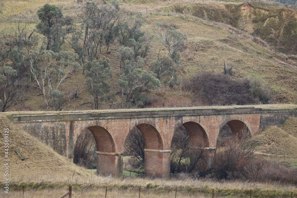 Old historic brick railway bridge in Australian countryside with green hills and eucalyptus trees in background on an overcast cold winter day.