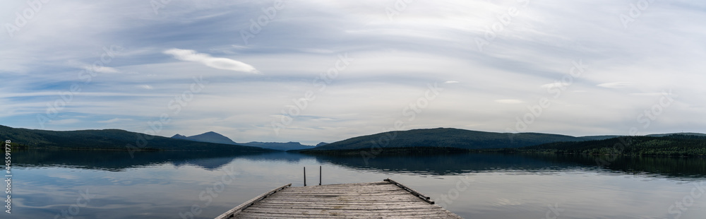 panorama of a calm lake with reflections of mountains and sky and a wooden dock in the foreground