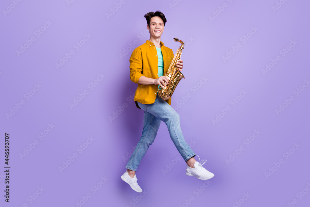 Full body profile side photo of young man happy positive smile jump up play saxophone isolated over violet color background Full body profile side photo of young man happy positive smile jump up play saxophone isolated over violet color background