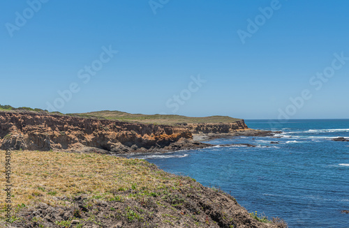 San Simeon, CA, USA - June 8, 2021: Pacific Ocean coastline north of town. Green plateau ending on red cliffs into deep blue water under light blue sky. Meadow and white surf.
