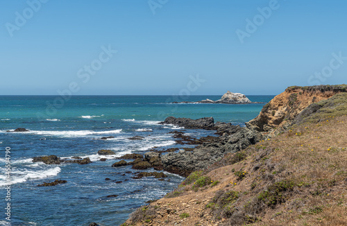San Simeon, CA, USA - June 8, 2021: Pacific Ocean coastline north of town. Piedras Blankas island in blue-azure ocean and low brown and black cliffs with white surf.