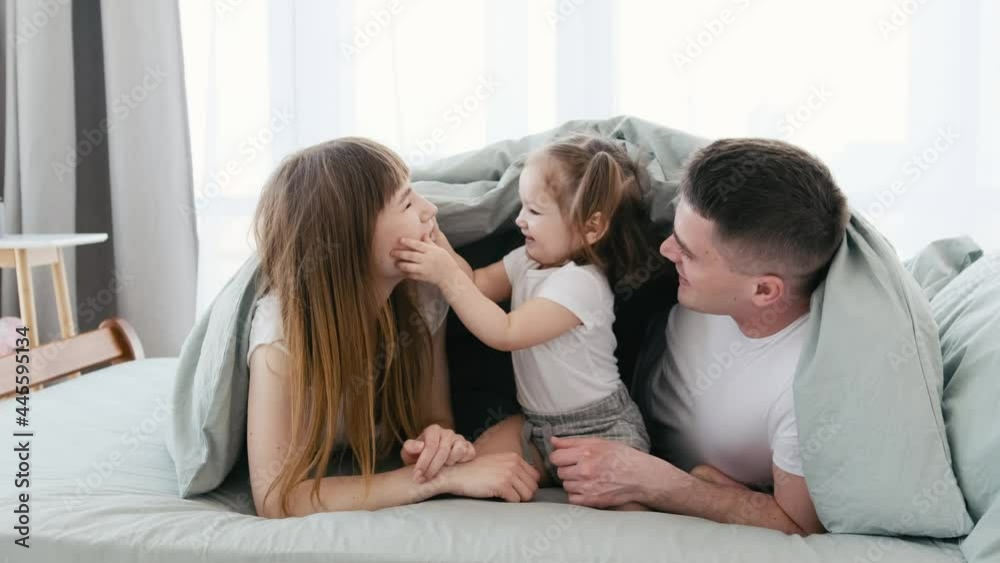 Young parents playing with little daughter and smiling under the blanket in the bed. Cute little girl kid touching mom and dad cheeks in the room with daylight. Father, mother and child. Young parents playing with little daughter and smiling under the blanket in the bed. Cute little girl kid touching mom and dad cheeks in the room with daylight. Father, mother and child.
