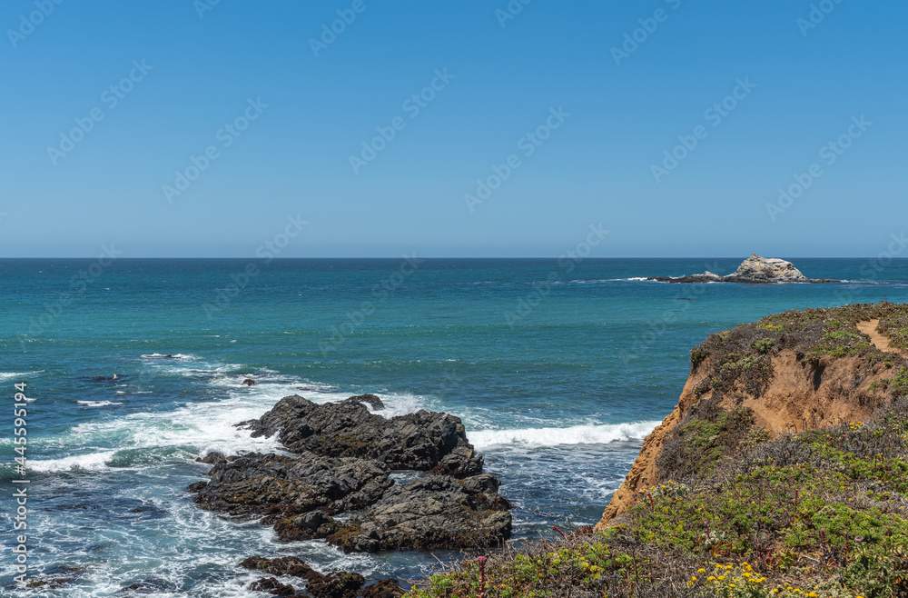 San Simeon, CA, USA - June 8, 2021: Pacific Ocean coastline north of town. Point Piedras Blancas with small white island and black rocks in blue azure water under light blue sky. 