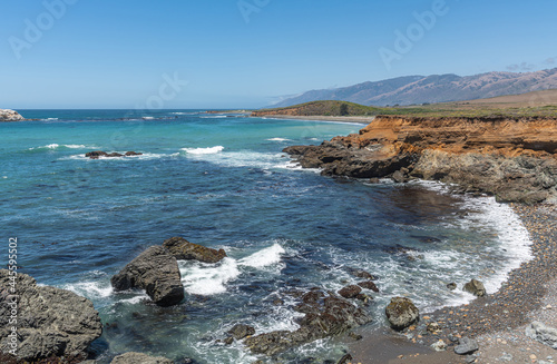 San Simeon, CA, USA - June 8, 2021: Pacific Ocean coastline north of town. Wide view on brown-red cliffs descend into blue-azure water under blue sky. Los Padres Mountain range on horizon.