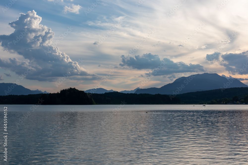 A panoramic view on the Lake Faak in Austria. The lake is surrounded by high Alpine peaks. The sun in slowly setting behind the mountains. Lots of clouds. Calm surface reflects the sunbeams. Serenity
