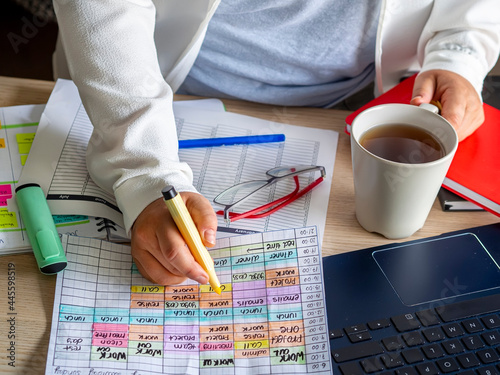 Woman hand highlighting on a weekly calendar and important appointment and holding a coffee cup. Work desk with schedule, laptop and paperwork. Time blocking for productivity and time management