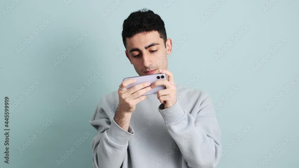 A happy brunette man is playing games on his mobile standing isolated over blue background in the studio A happy brunette man is playing games on his mobile standing isolated over blue background in the studio