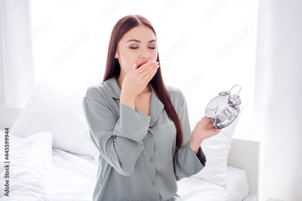 Photo of charming tired young lady grey sleepwear sitting bed arm cover lips yawning holding clock indoors room home house Photo of charming tired young lady grey sleepwear sitting bed arm cover lips yawning holding clock indoors room home house
