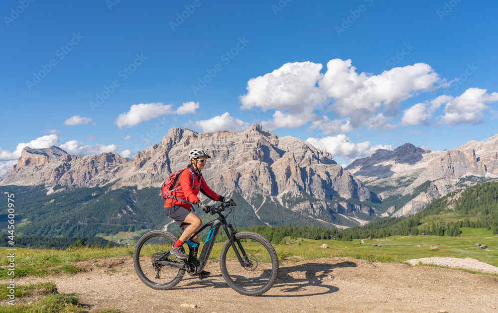 nice and active senior woman riding her electric mountain bike on the Pralongia Plateau in the Alta Badia Dolomites with awesome Sasso die Santa Cruce summit in Backg, South Tirol and Trentino, Italy