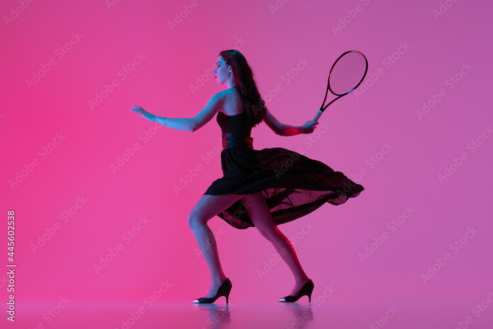 High-fashion styled young woman in black evening gown, dress playing tennis isolated over pink neon background. Concept of fashion and sport