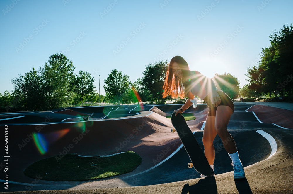 Asian skater girl having fun when she is skateboarding. 
She is wearing a pink t-shirt, black pants, white socks, and white sneakers. 
She is at the skatepark and as you can see the sunset is awesome! - obrazy, fototapety, plakaty 