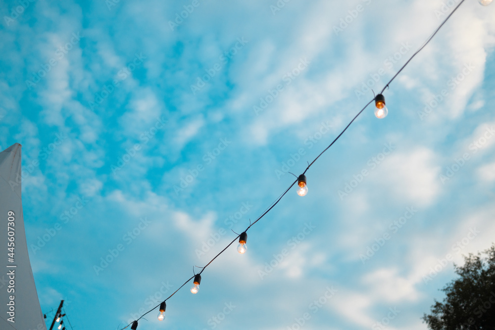 Fashion decoration string lights hanging in restaurant or cafe in the garden at summer party. Outdoor electric lamps. Bright blue sky
