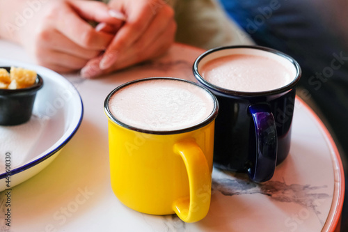Two colored mugs with a hot drink and foam stand on the table against the background of women s hands.