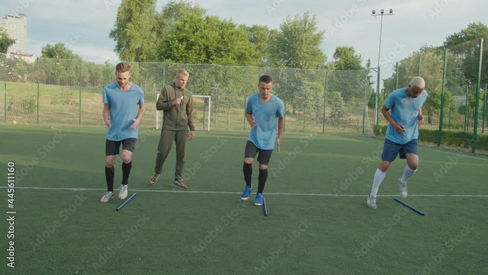 Team of diverse multiracial soccer players in football uniform performing jog on the spot exercise outdoors at sunrise, while young trainer urging sportsmen to move faster, looking on hand watch Team of diverse multiracial soccer players in football uniform performing jog on the spot exercise outdoors at sunrise, while young trainer urging sportsmen to move faster, looking on hand watch