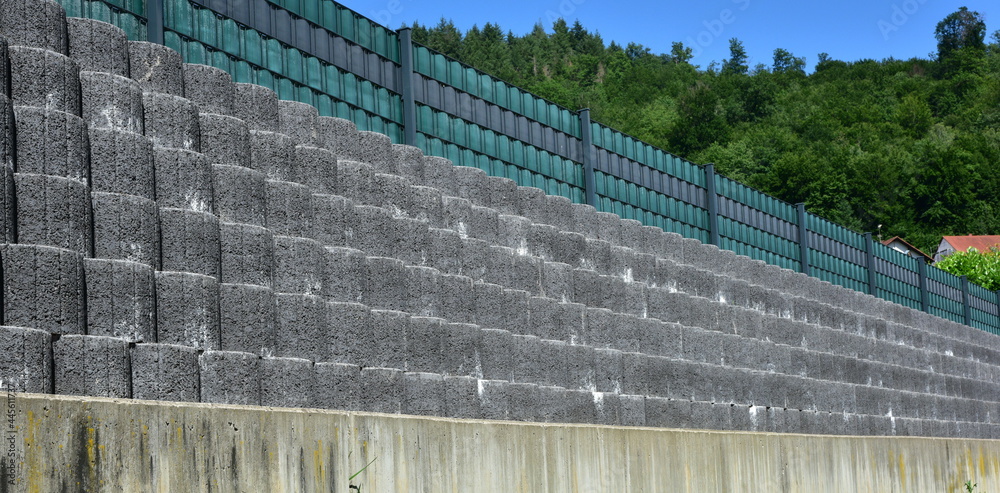 Stützmauer aus grauen runden Pflanzsteinen zur Hangsicherung und Abgrenzung eines Wohngrundstücks gegen die tiefer liegende Straße - obrazy, fototapety, plakaty 