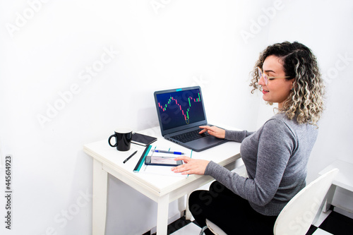 young woman working from home analyzing financial charts on computer and cell phone