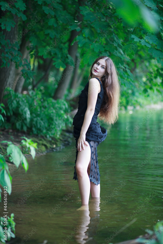 A beautiful young romantic girl with long flowing hair in a black long dress stands in the green water of a summer river or lake. A beautiful young romantic girl with long flowing hair in a black long dress stands in the green water of a summer river or lake.