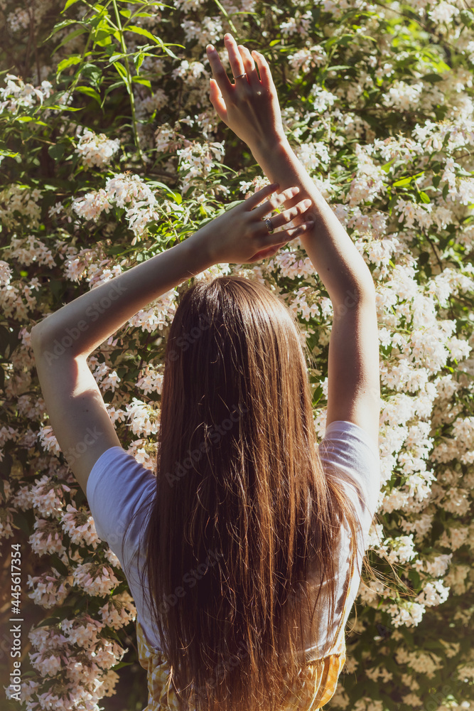 Fototapeta premium Beautiful slender brunette girl raised her hands up and posing back against the background of flowering bushes on a sunny day