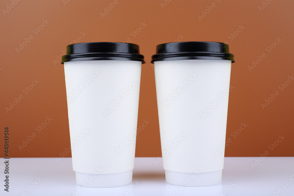Close up of two white cardboard cup of coffees on white table against brown background. Concept of hot drinks for take away Close up of two white cardboard cup of coffees on white table against brown background. Concept of hot drinks for take away